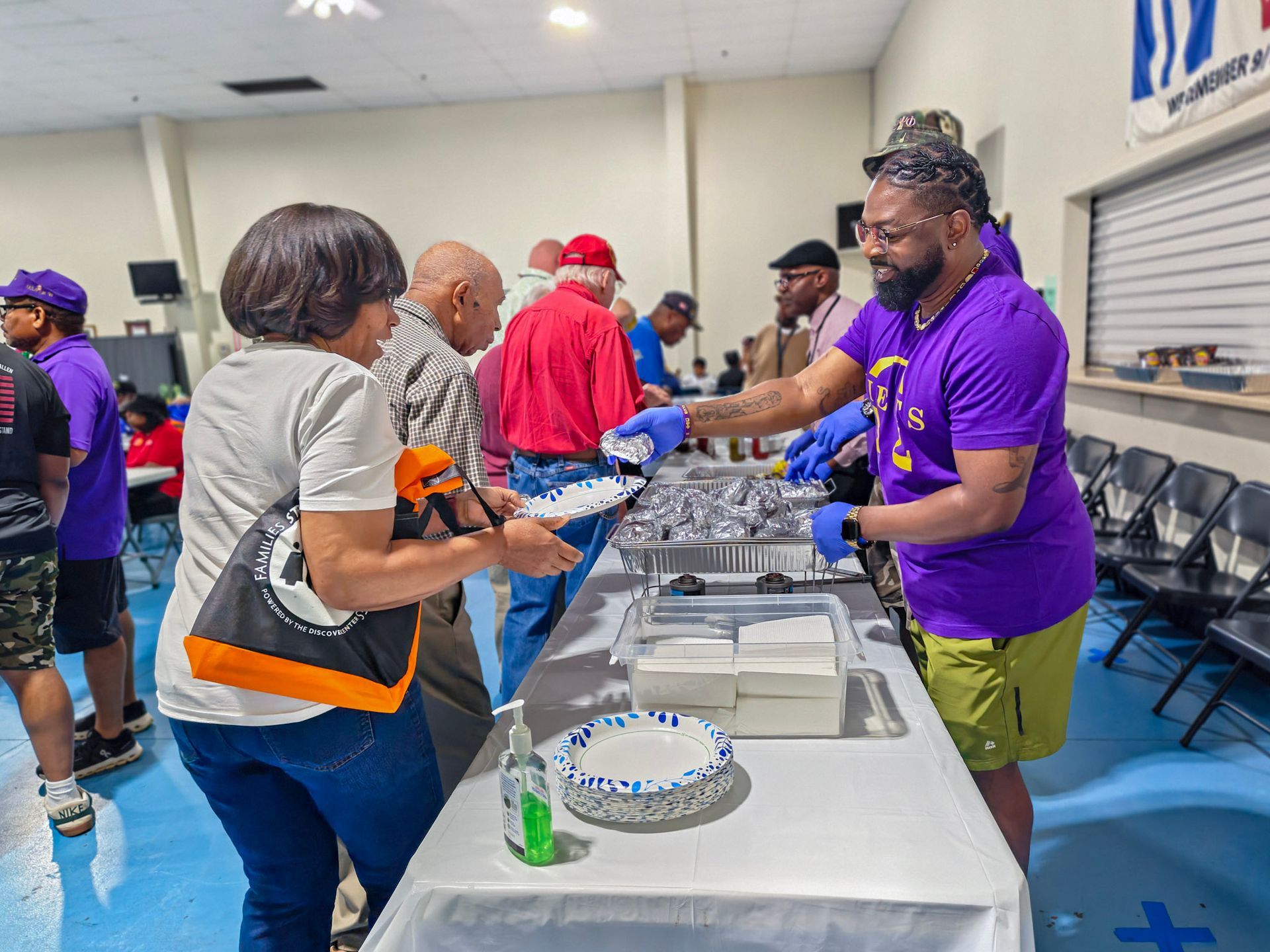 A volunteer wearing a purple shirt serves food on a paper plate to a person in a community hall setting.