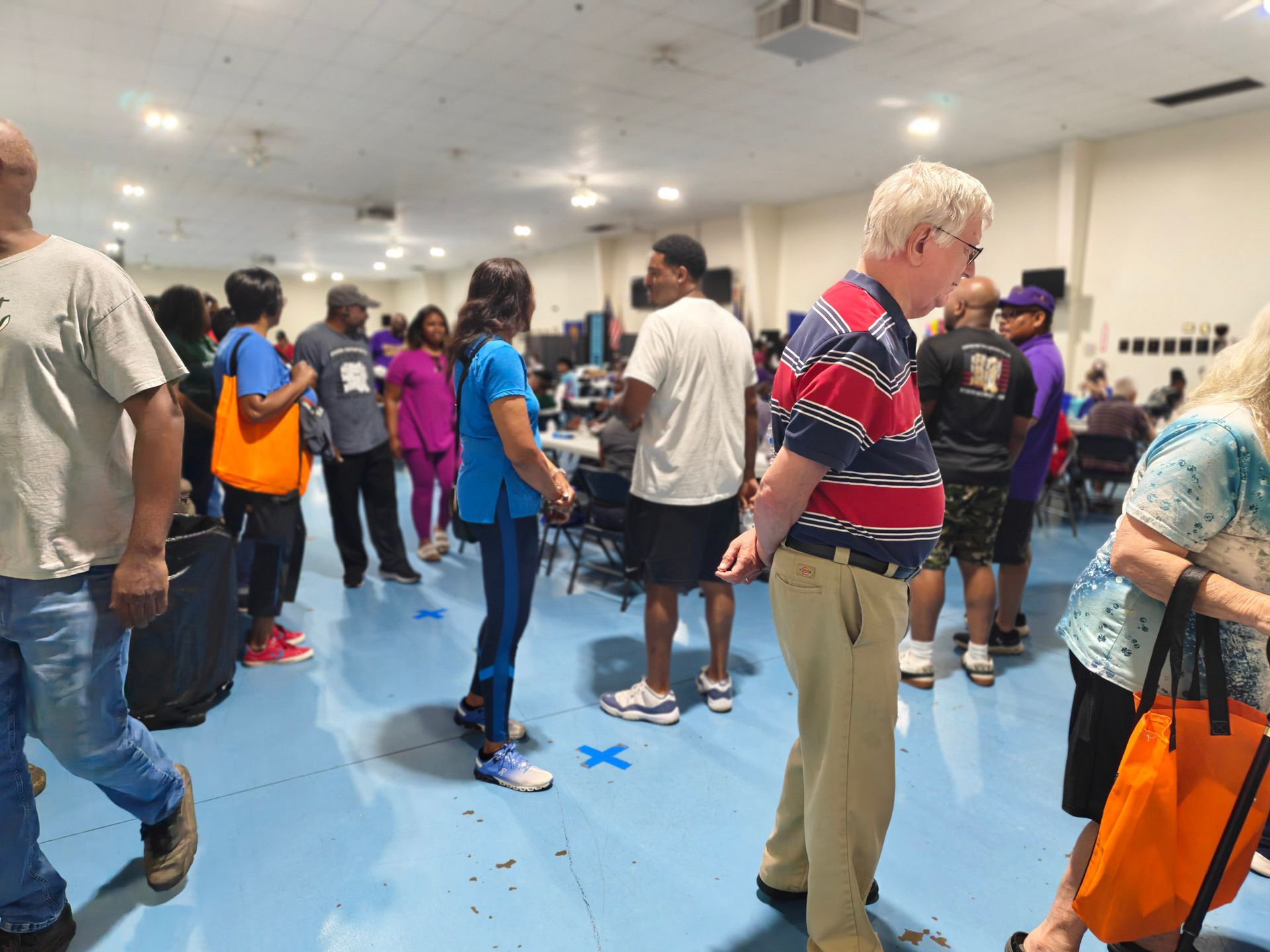People stand in a line across a large hall with blue flooring, some carrying bags, appearing to wait for an event.