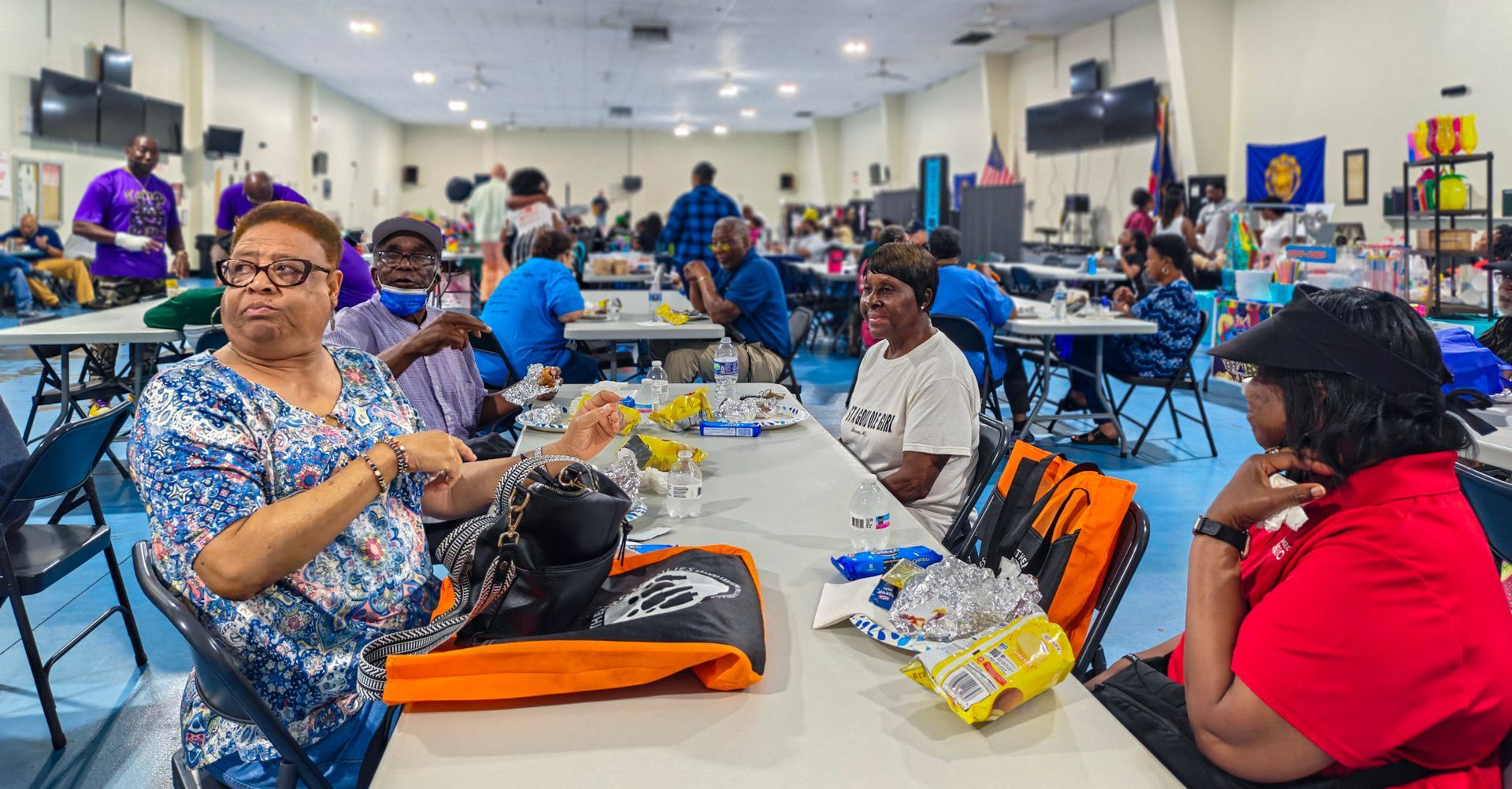 People seated at tables in a large hall for a social gathering, with food items on the tables and others in the background.