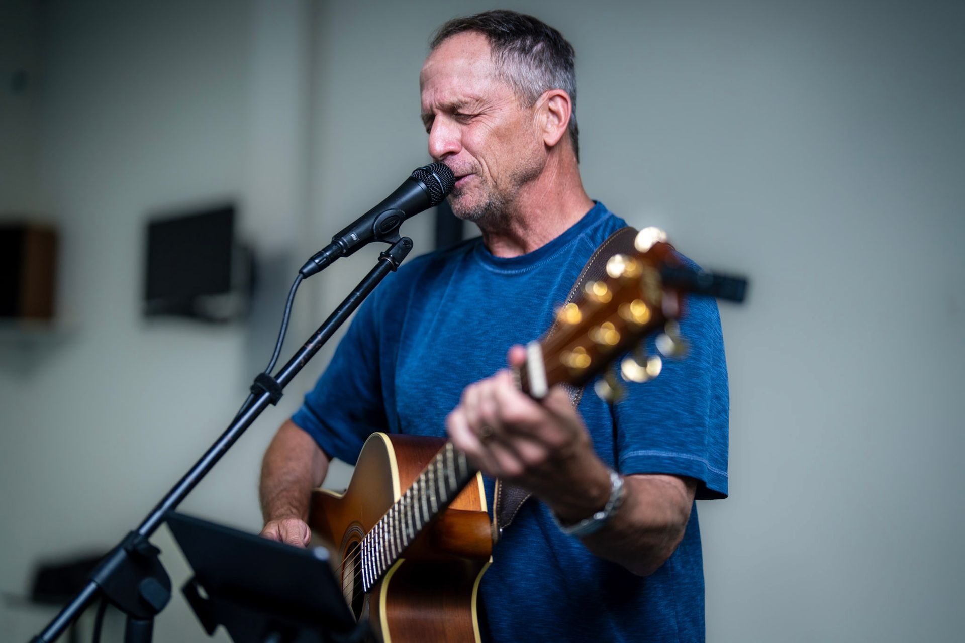 A person in a blue shirt plays an acoustic guitar and sings into a microphone on a stand.