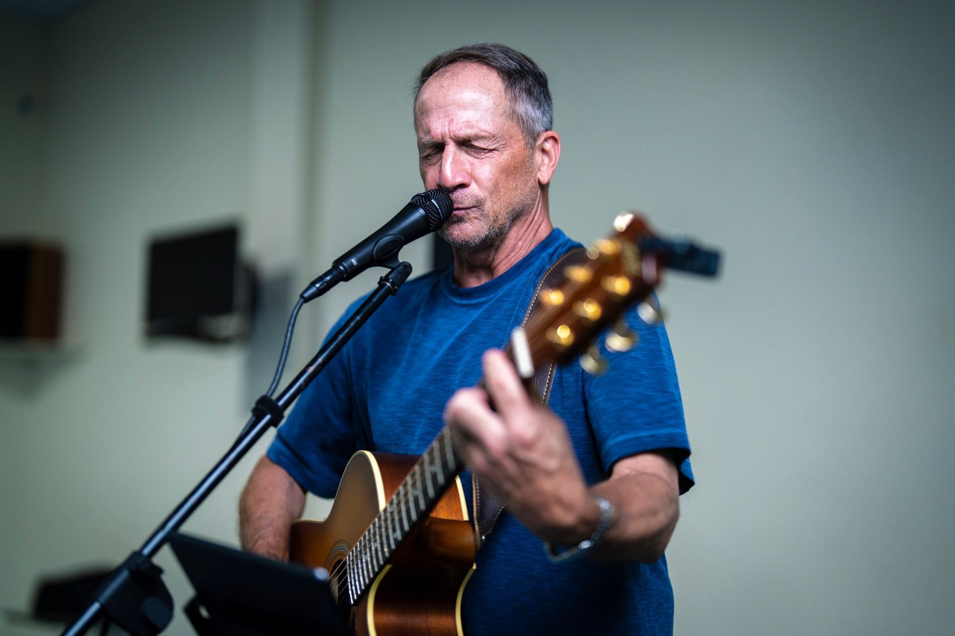 A focused performer plays an acoustic guitar and sings into a microphone against a plain wall.