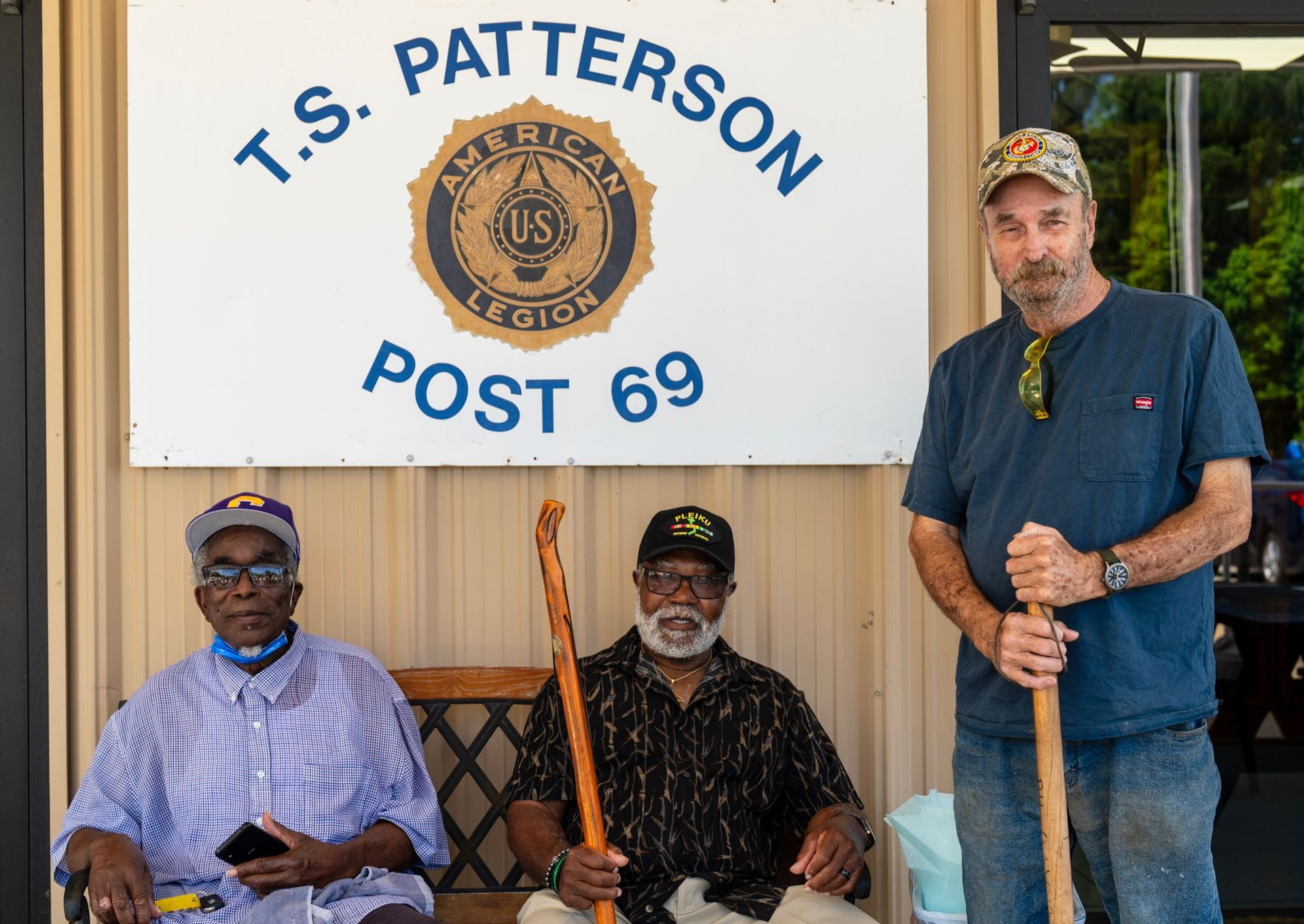 Three people stand or sit in front of an American Legion Post 69 sign on a building wall.