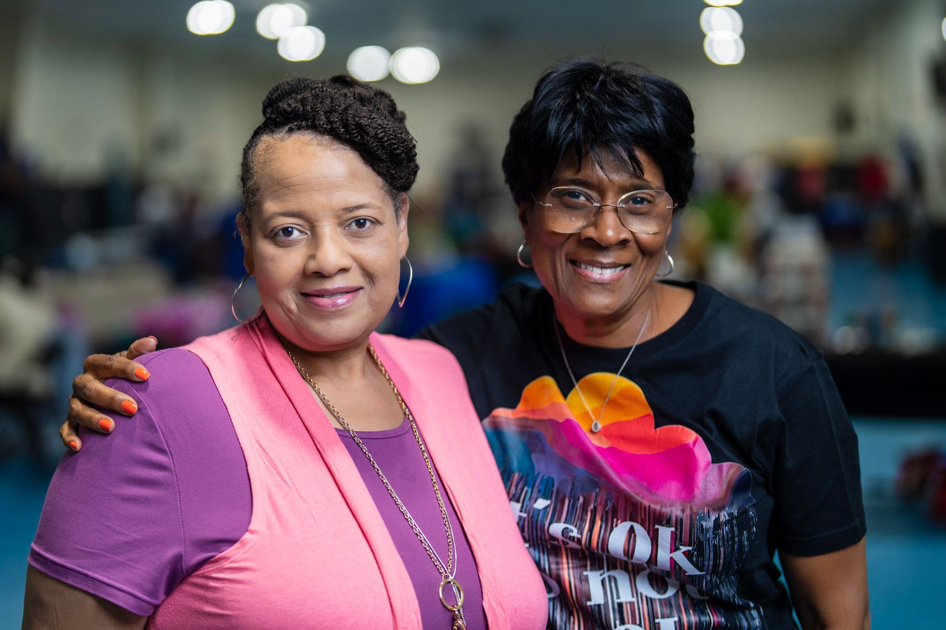 Two smiling people standing close together indoors, one wearing a pink vest over a purple shirt and the other in black.
