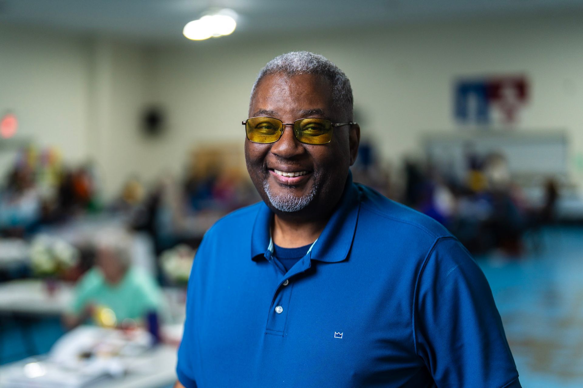 Smiling person in a blue polo shirt and yellow-tinted glasses in a brightly lit community room with blurred tables.