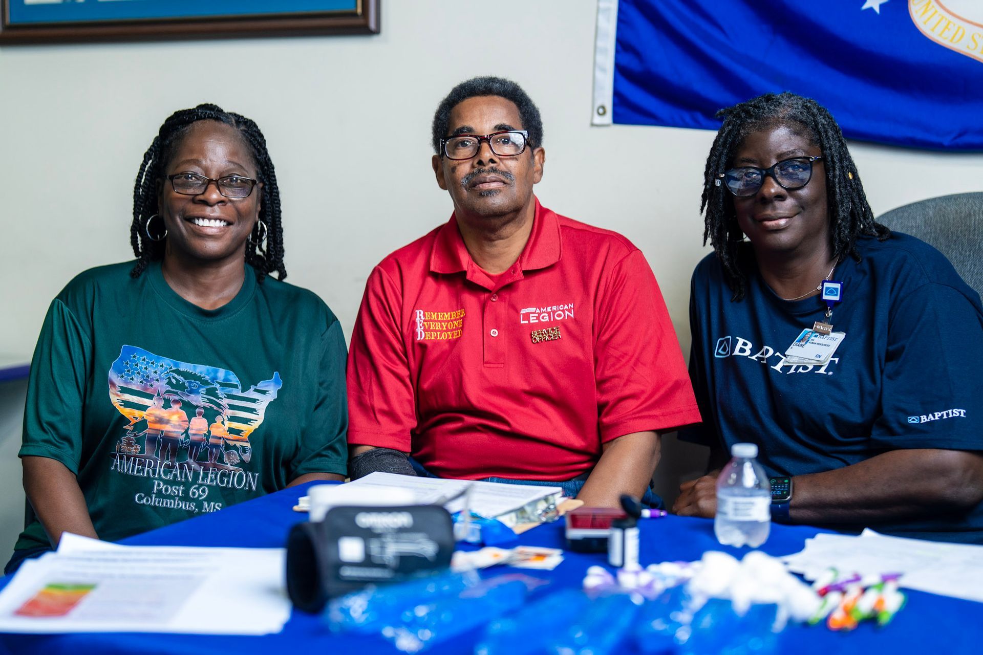 Three people sit behind a blue table with medical supplies, smiling at the camera in a room with a flag.