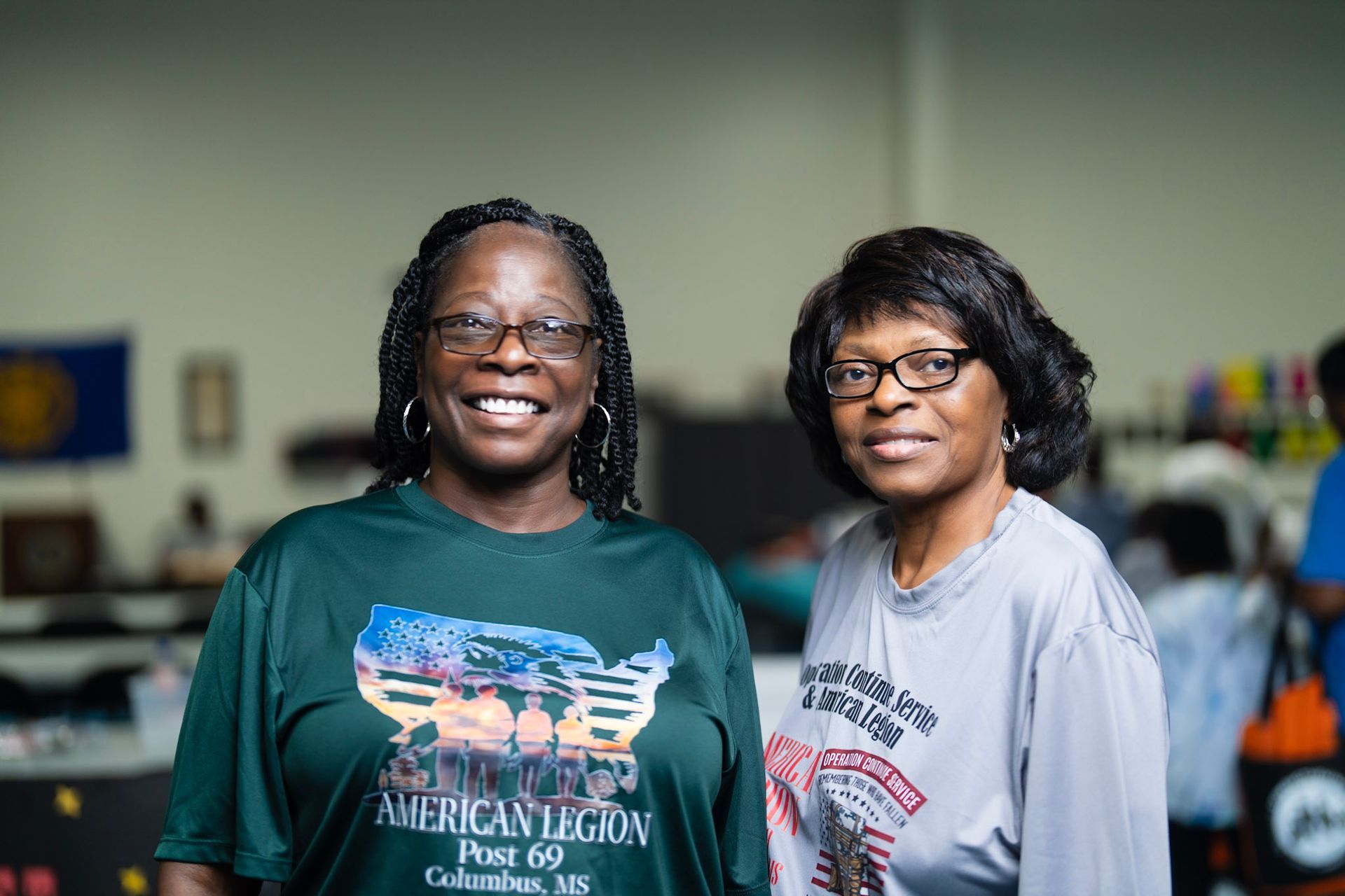 Two women stand indoors wearing American Legion T-shirts, one smiling and one looking toward the camera.