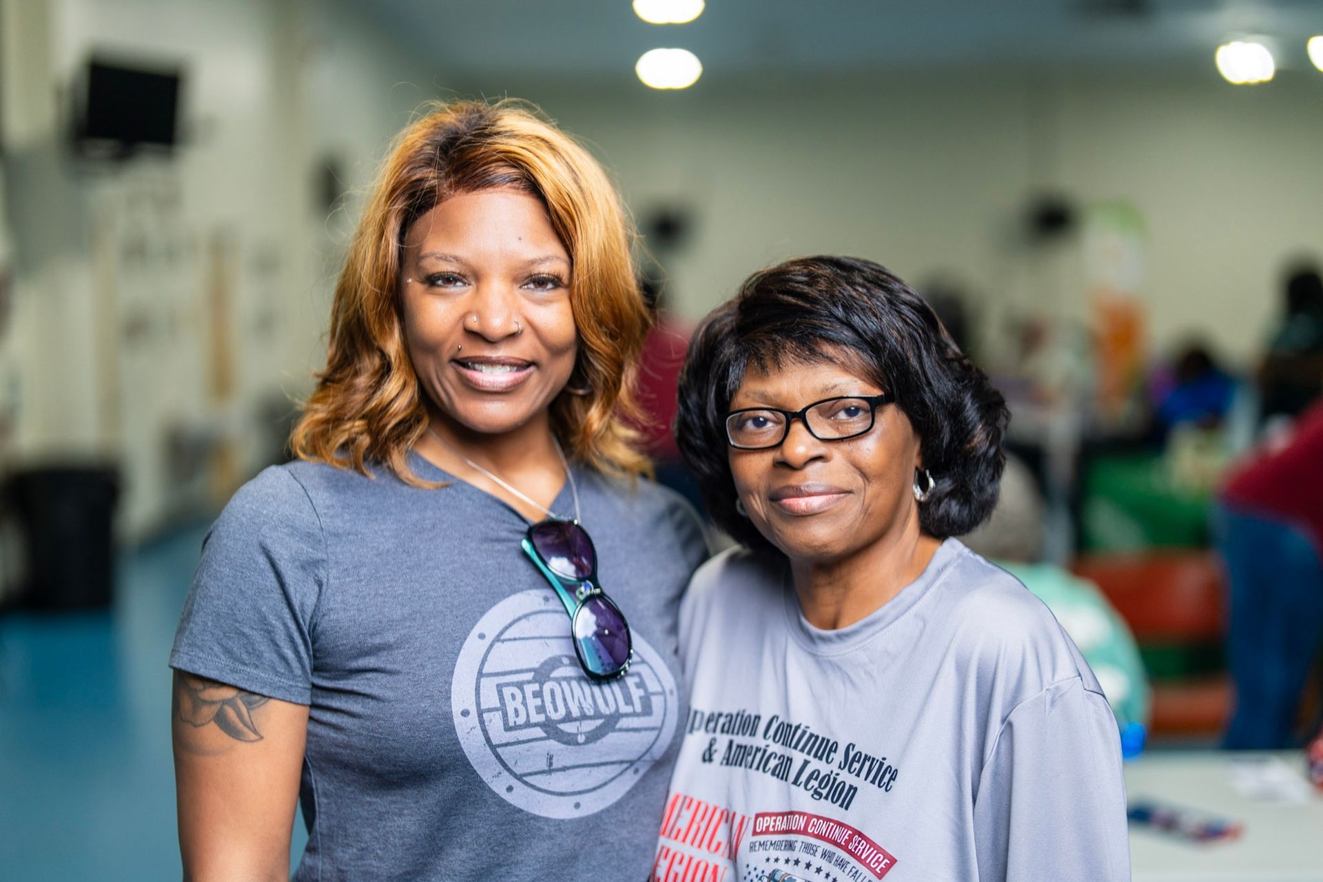 Two smiling people stand closely side-by-side in a well-lit community room, wearing casual t-shirts.