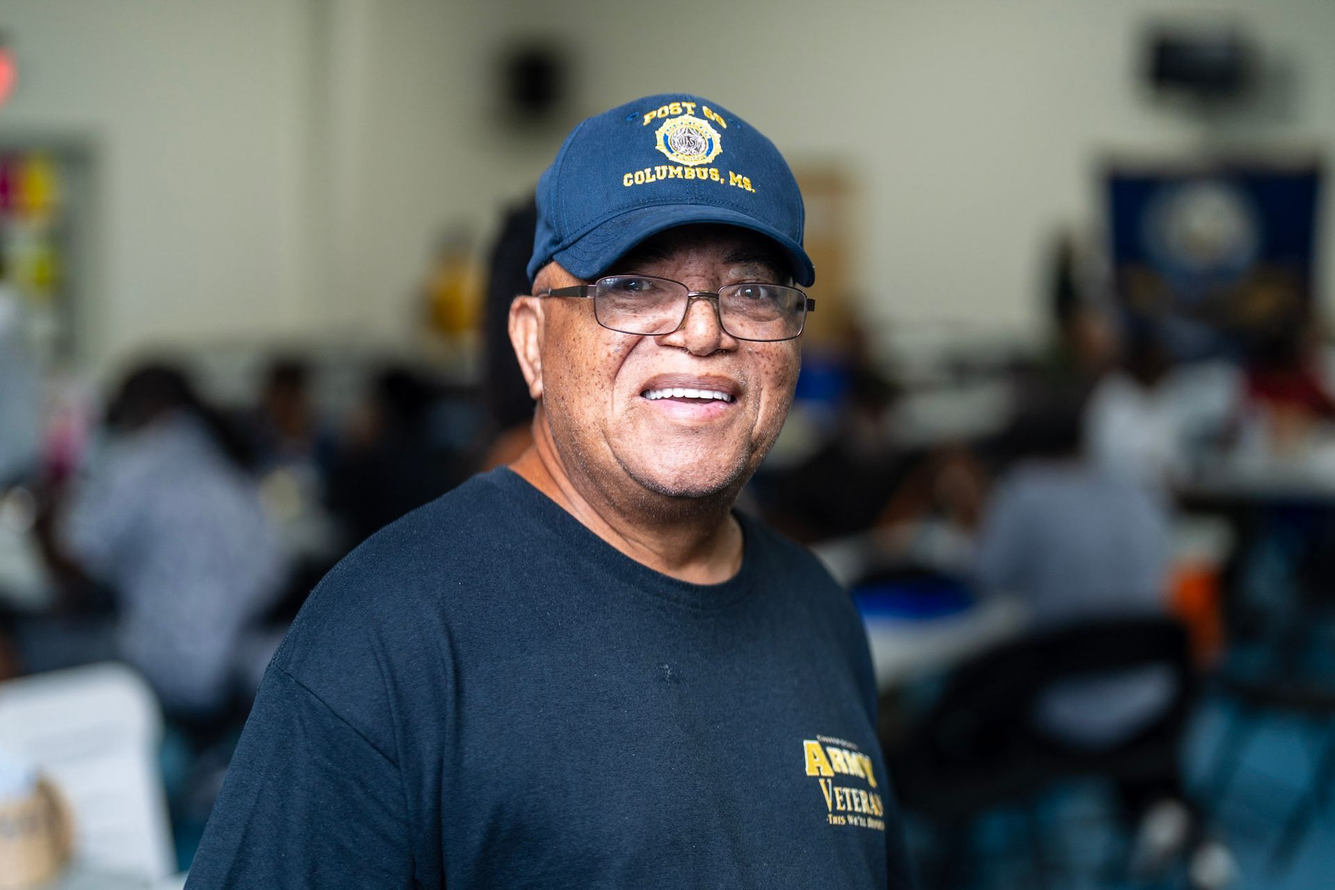 A smiling person wearing a dark blue cap and matching t-shirt in an indoor setting with blurred tables and chairs.