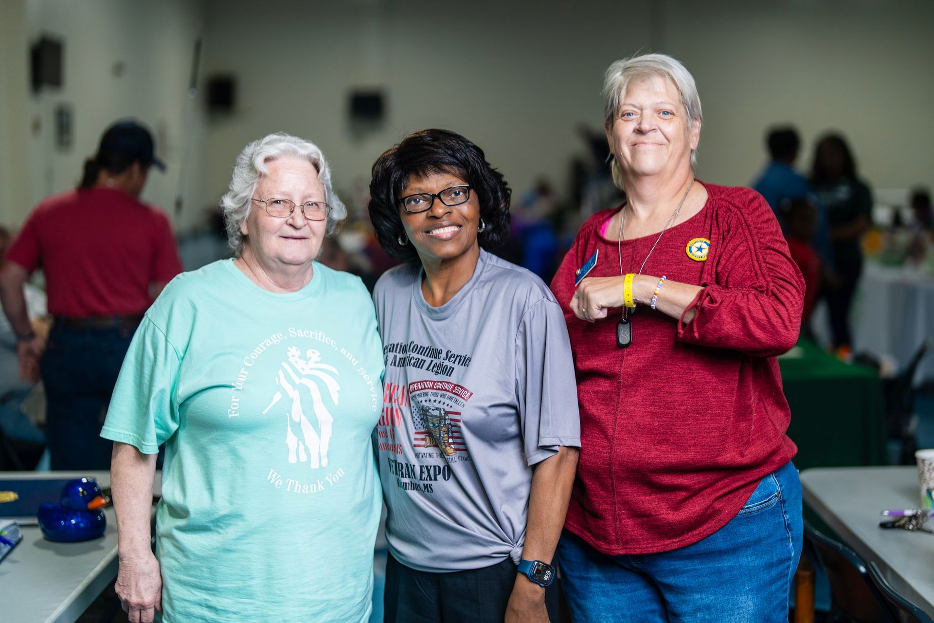 Three people stand side-by-side indoors, smiling at the camera. They wear casual clothing in a community hall setting.