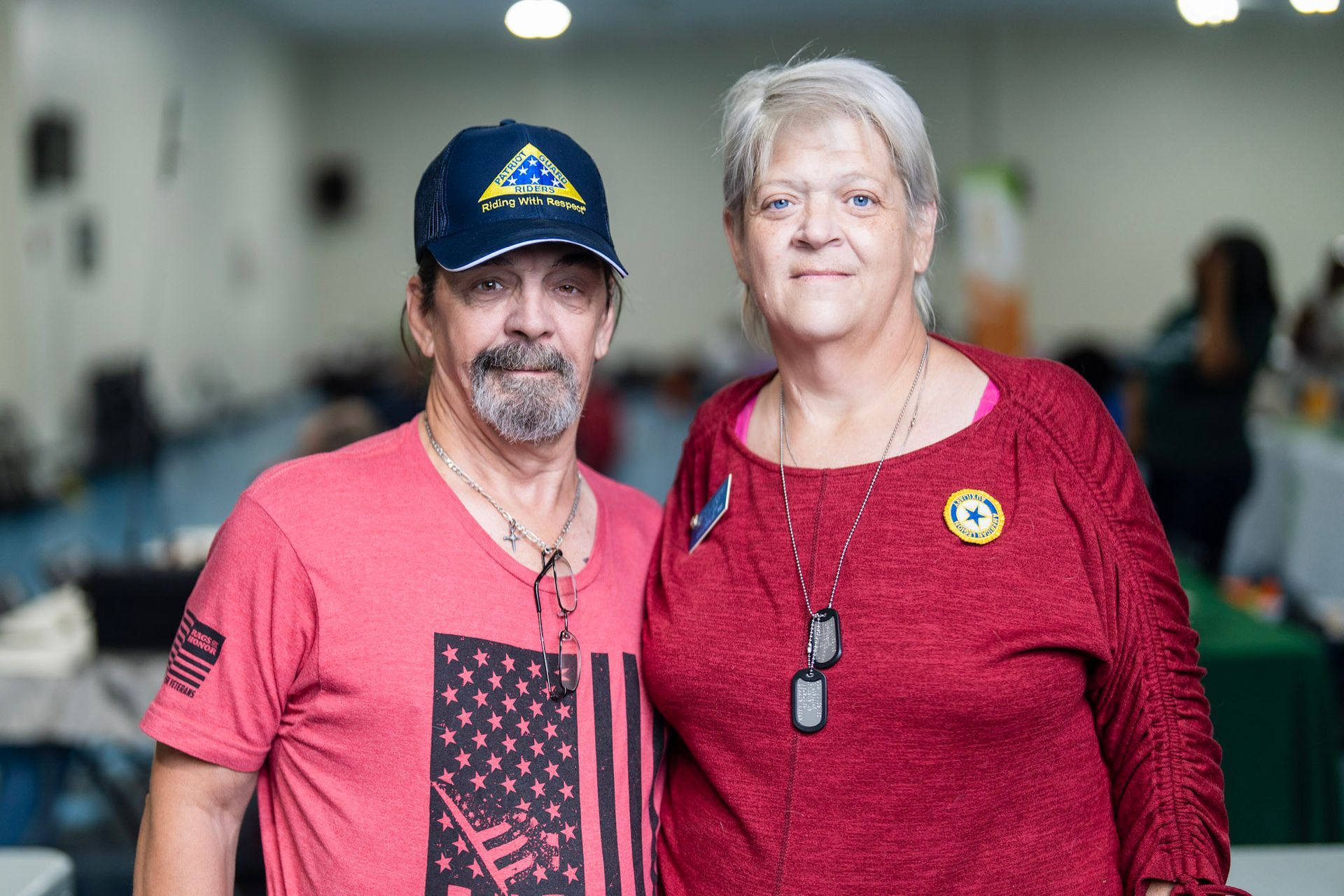 Two people pose for a photo indoors; the person on the left wears a baseball cap and red shirt, the right a red top.