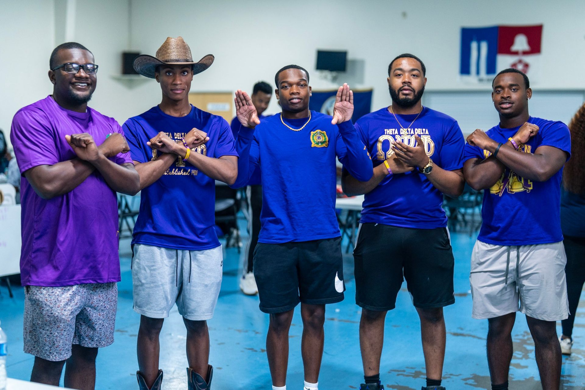 Five people stand in a line indoors wearing blue or purple shirts and shorts, holding hand signals while facing the camera.