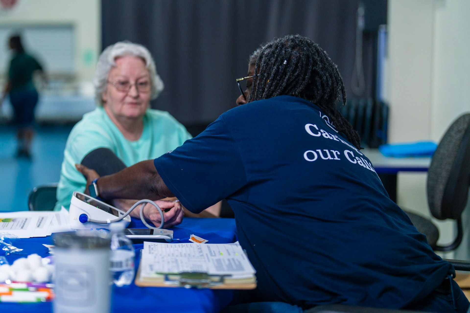 A person in a blue shirt checks a patient's blood pressure at a table in a brightly lit indoor space.