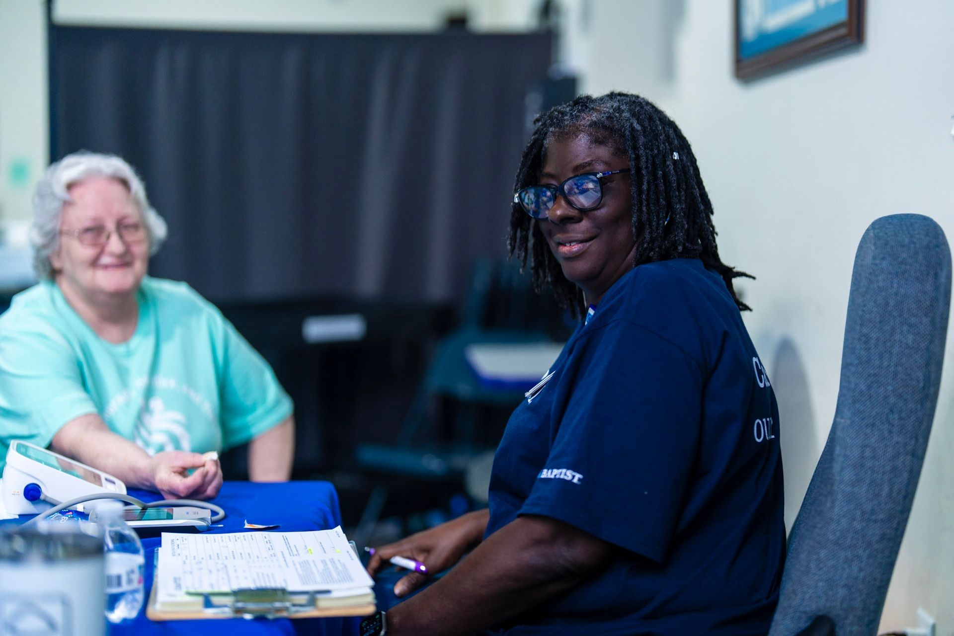 A person in a blue shirt smiling at a desk while a second person sits nearby with medical equipment on a blue table.