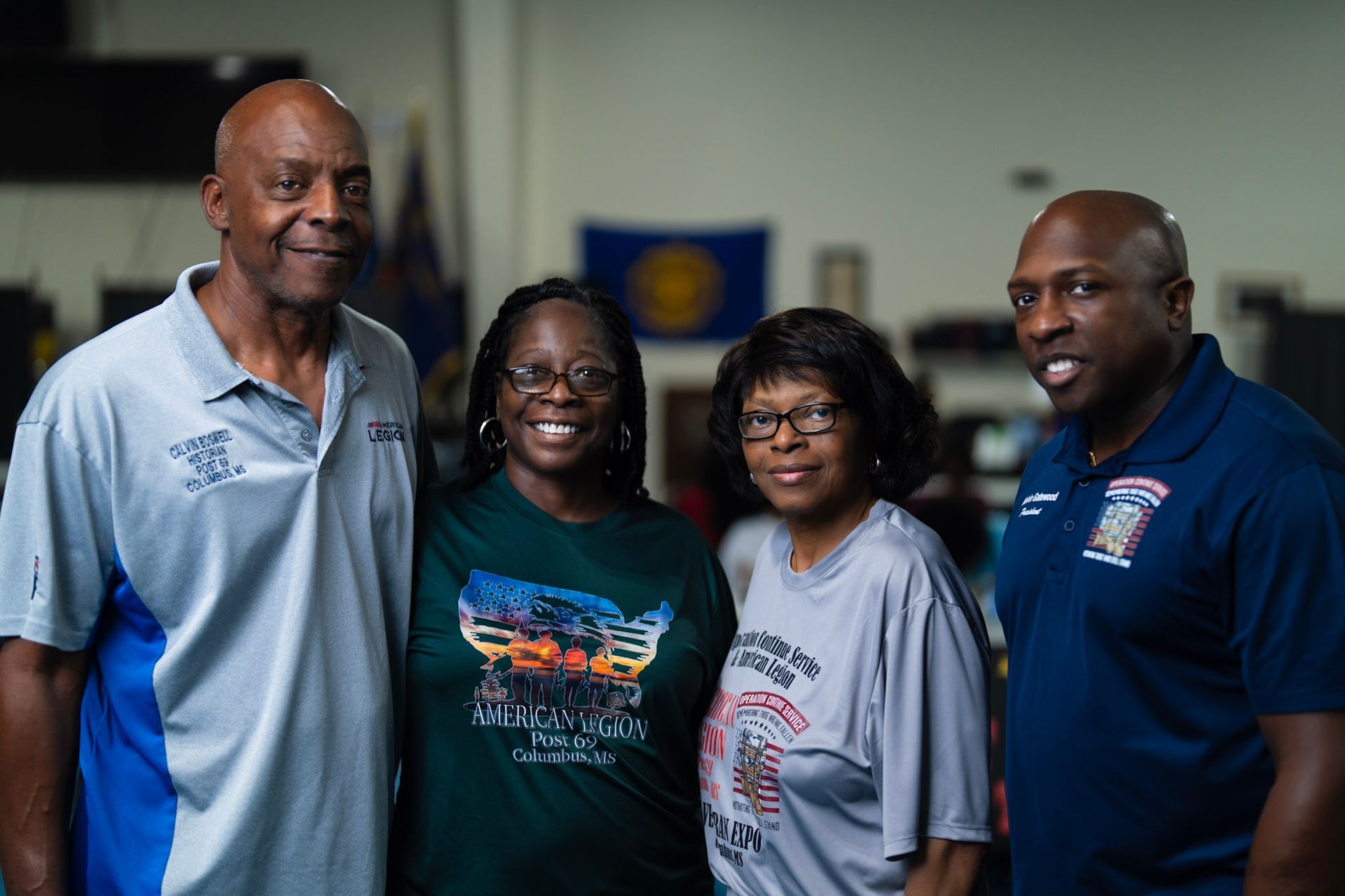 Four people pose smiling for a photo in an indoor room with a flag on the wall.