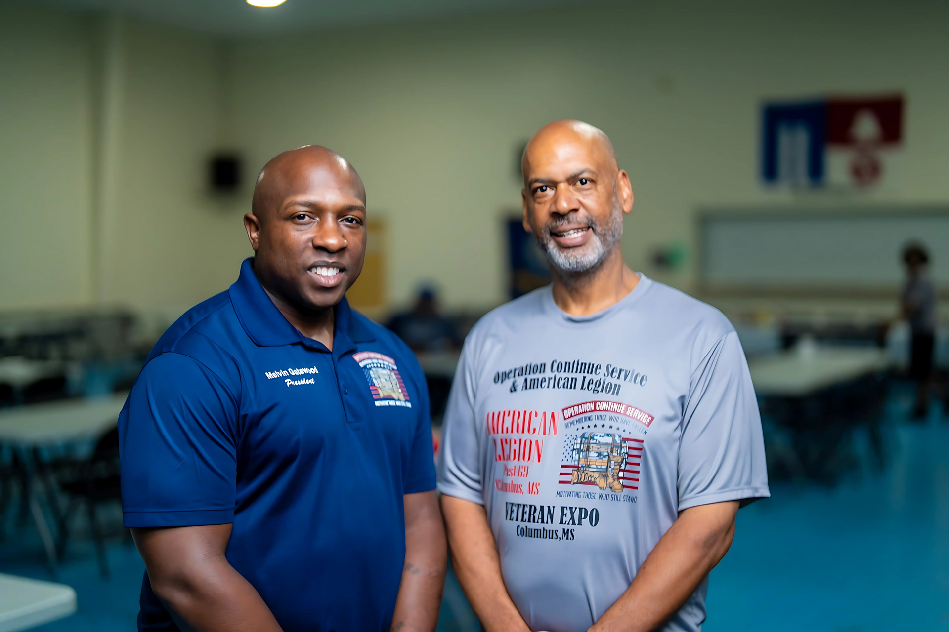 Two individuals stand side-by-side in a large, brightly lit room with tables, one wearing a navy shirt and one a grey shirt.