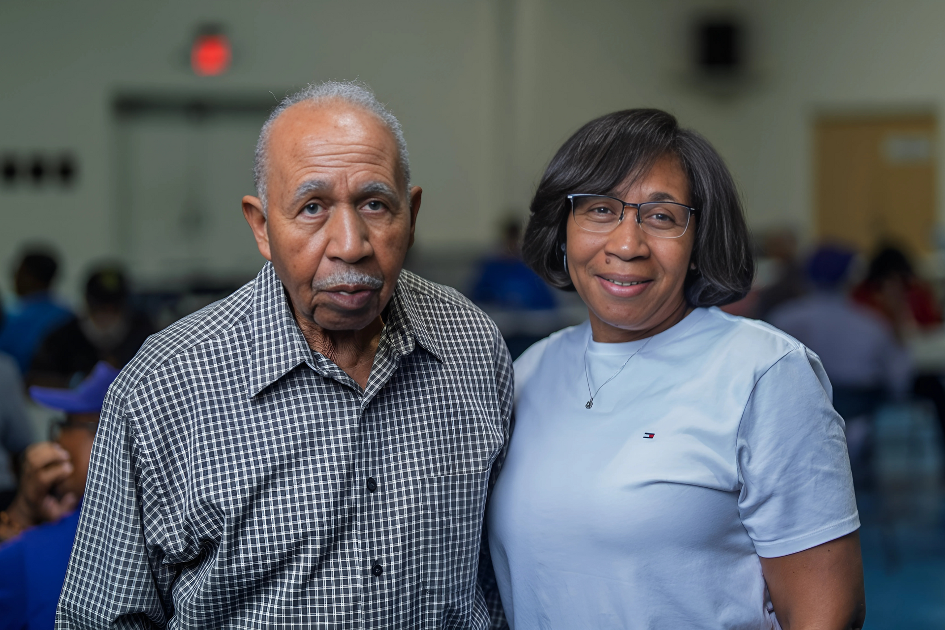 An older person and a middle-aged person standing side-by-side, smiling, in a bright, indoor public space.
