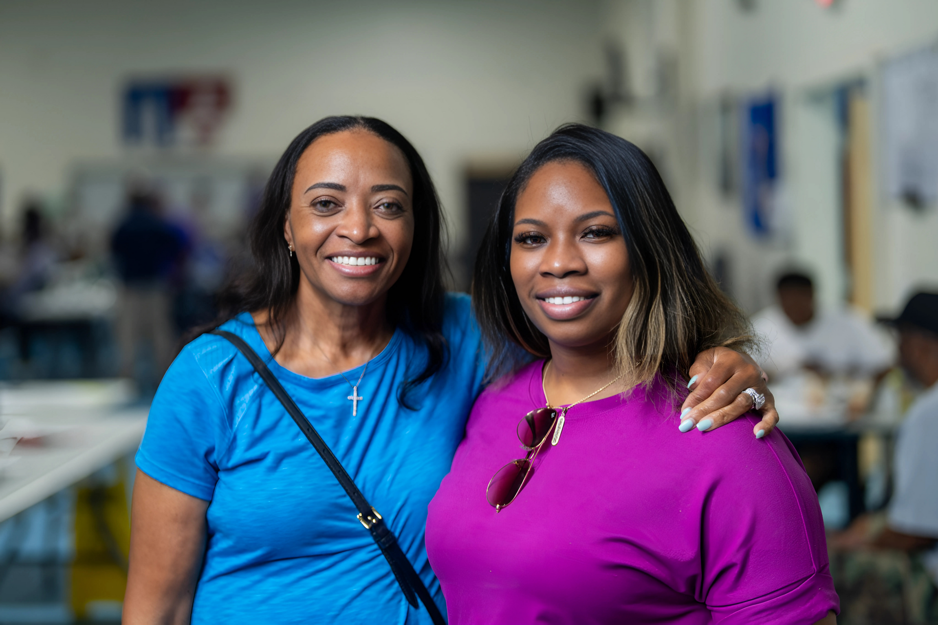 Two smiling people pose together indoors; one wears a blue top with a shoulder bag, the other a pink top with sunglasses.O