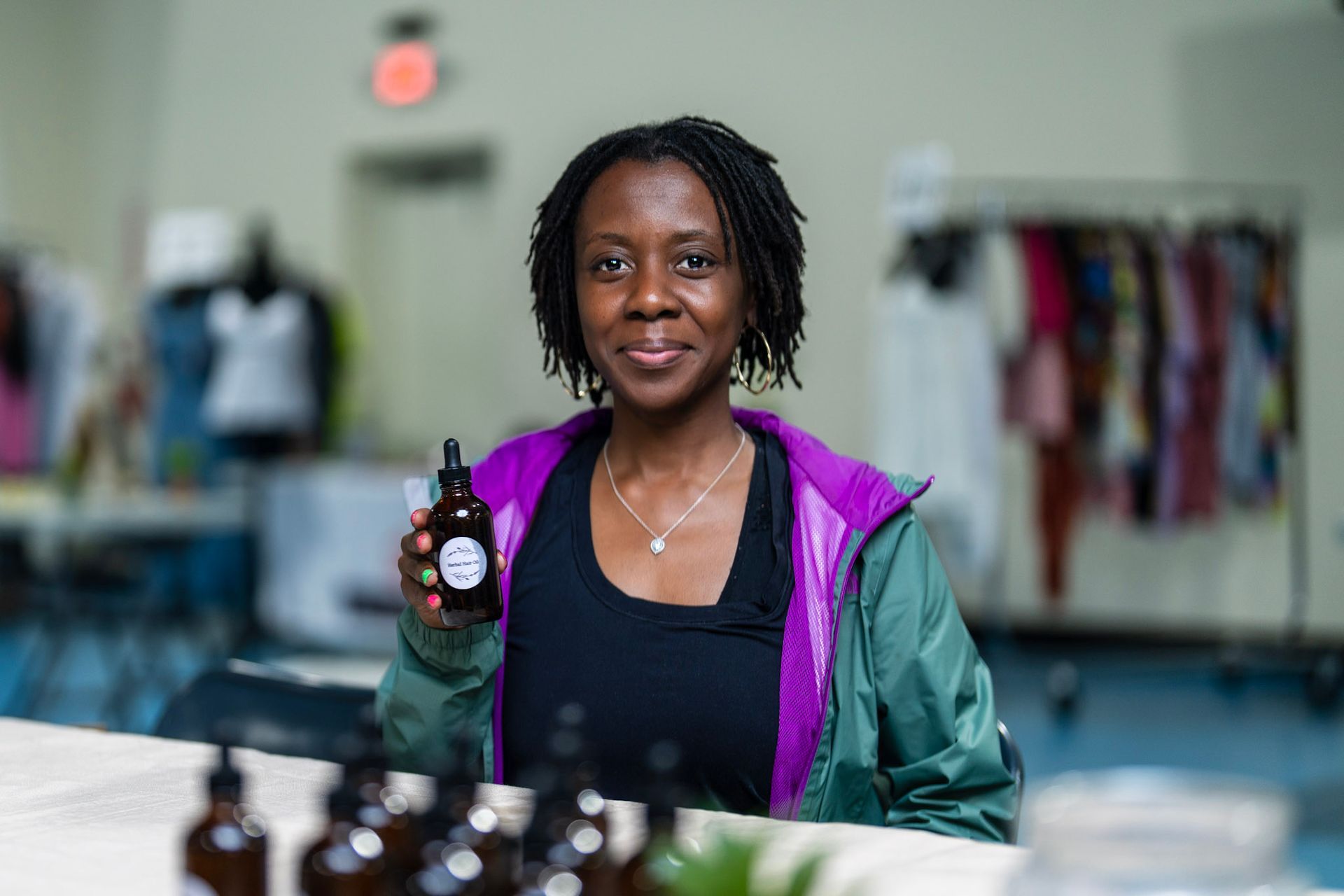 A person in a purple and green jacket holds a brown dropper bottle in a room with clothing racks.