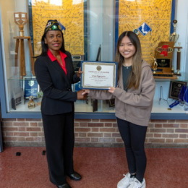 A person in a suit and cap presents a certificate to a smiling student in a hoodie in front of a trophy display case.