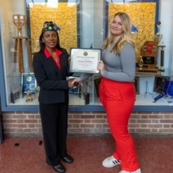 Two people stand in front of a display case; one hands a certificate to the other. Both are smiling.