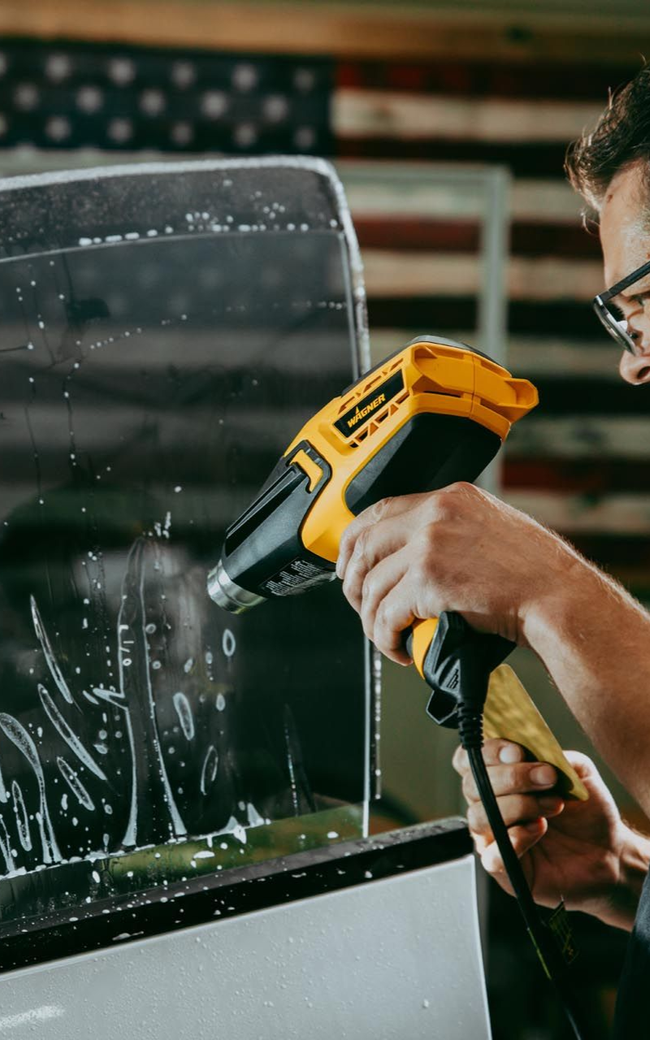 Person using a heat gun to apply tint to a car window. In a garage setting with an American flag.