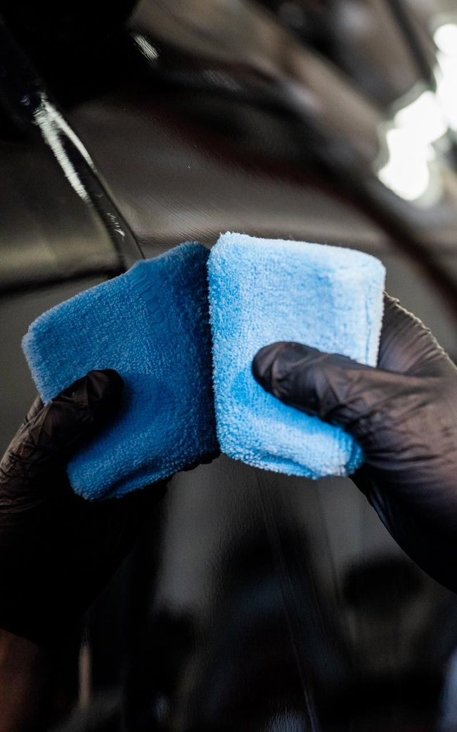 Gloves holding two blue microfiber cloths, wet, in an automotive setting.