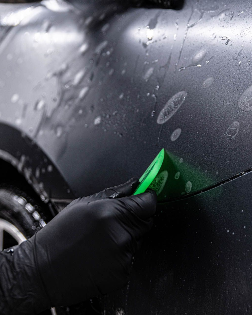 Gloved hand using a green squeegee on a wet, matte black car surface.