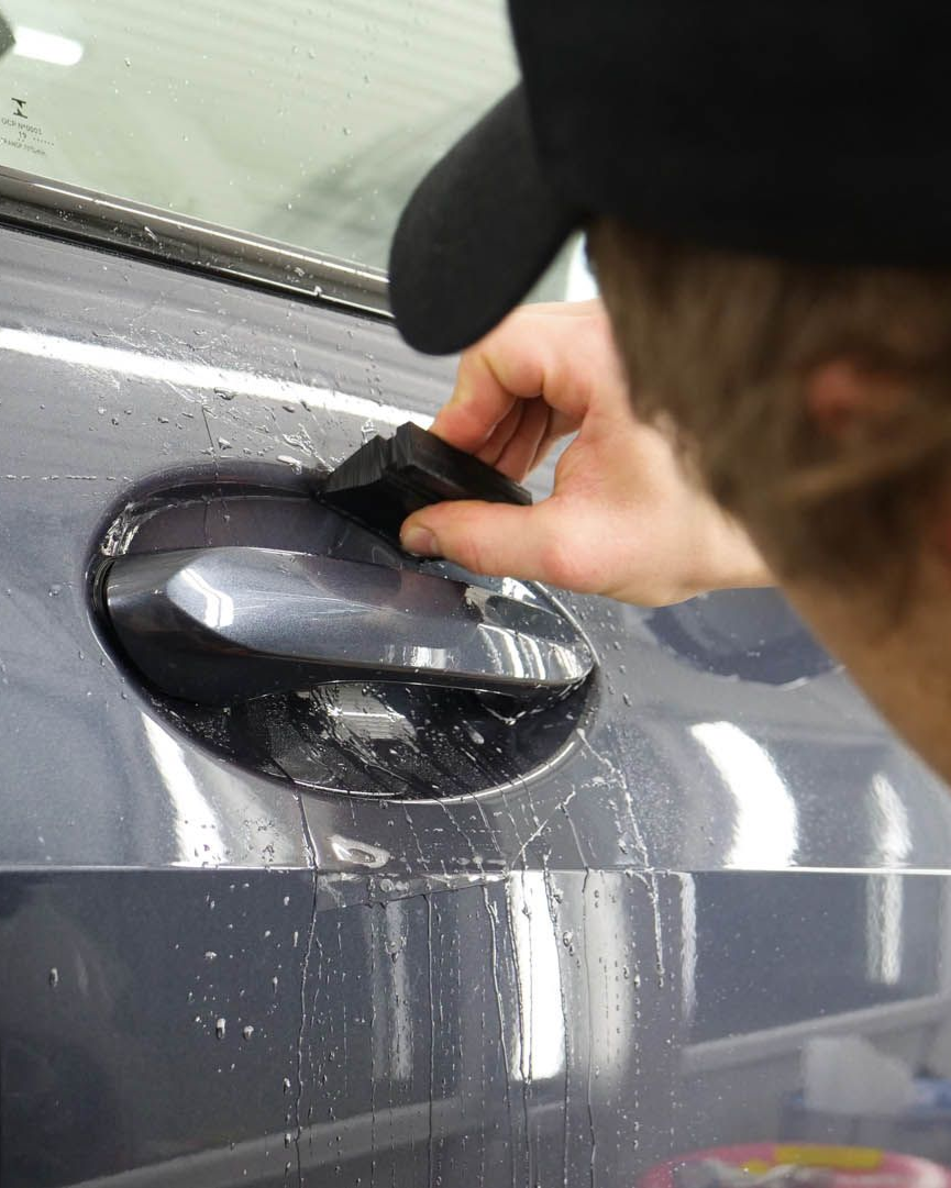 Person using a tool to apply a liquid to a car door handle. The car is blue.