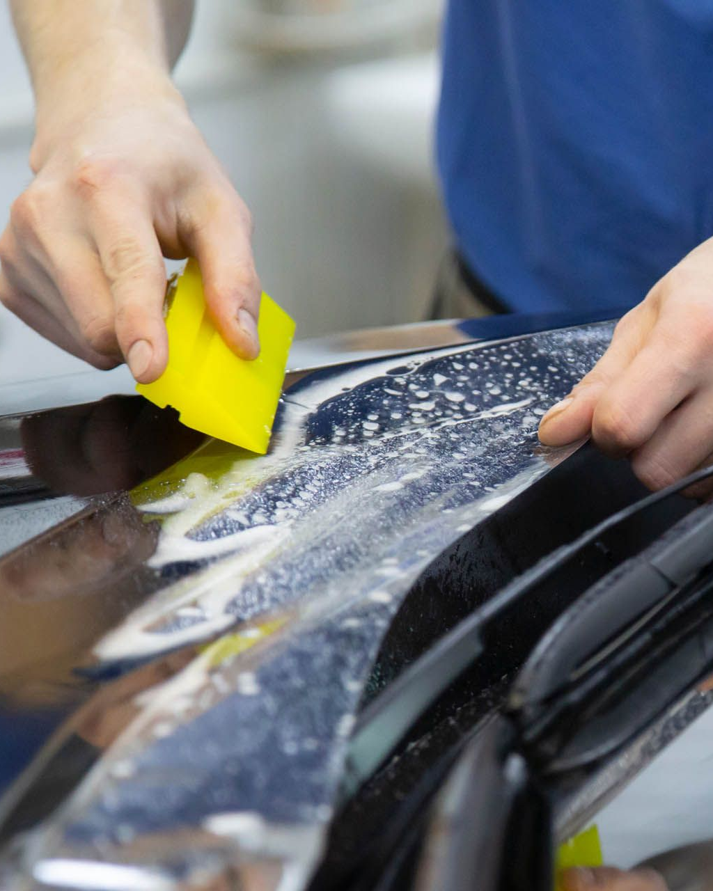 Person using a yellow squeegee to apply film to a dark blue car.