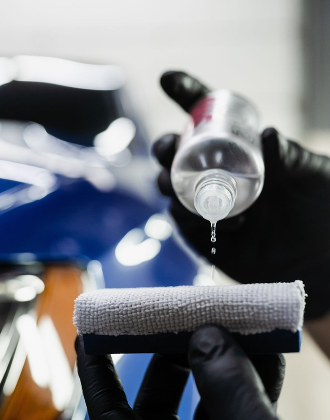 Person wearing black gloves polishing a silver car with a rotary buffer, outdoors.
