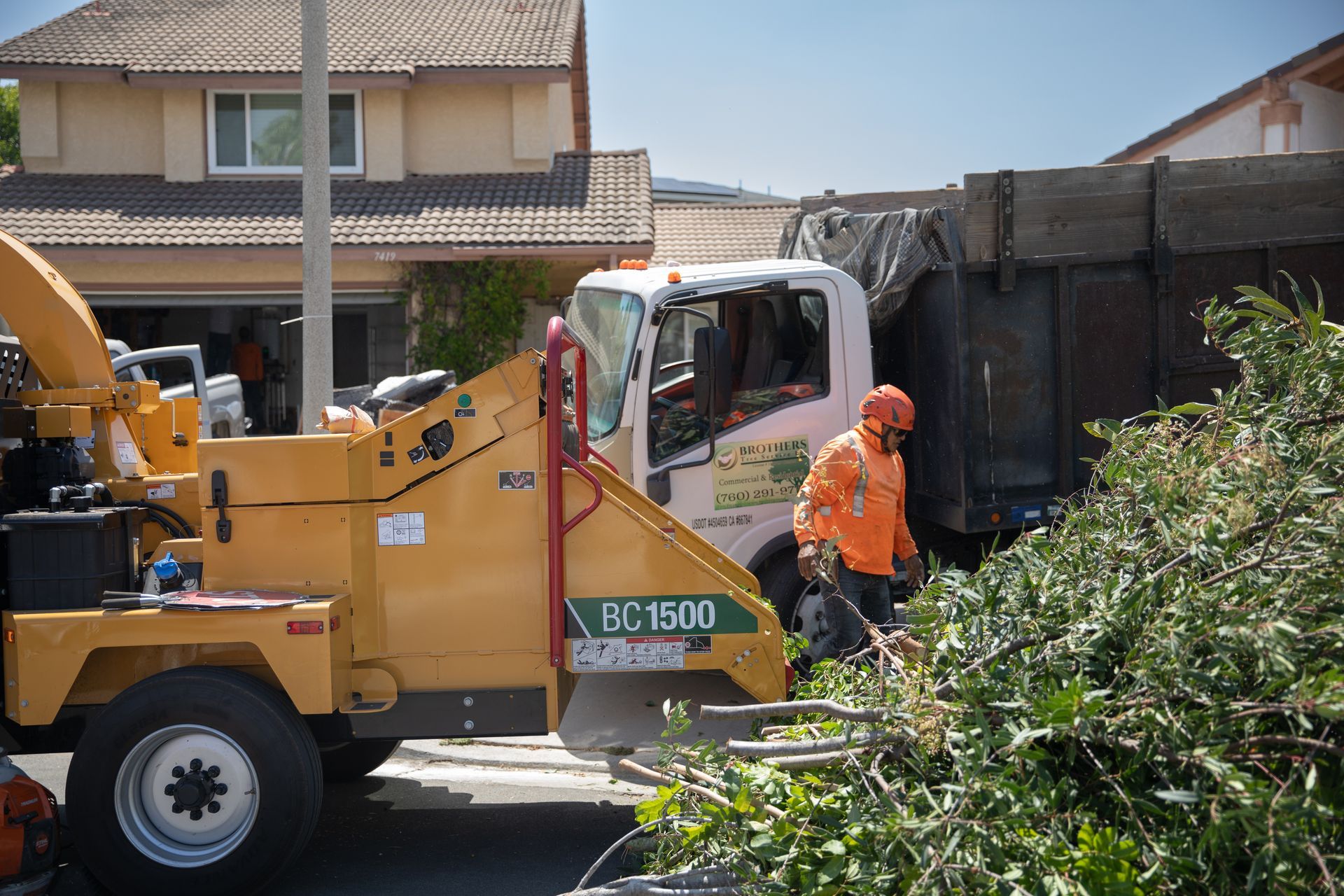 Orange wood chipper truck with worker in safety gear clearing debris beside a suburban house