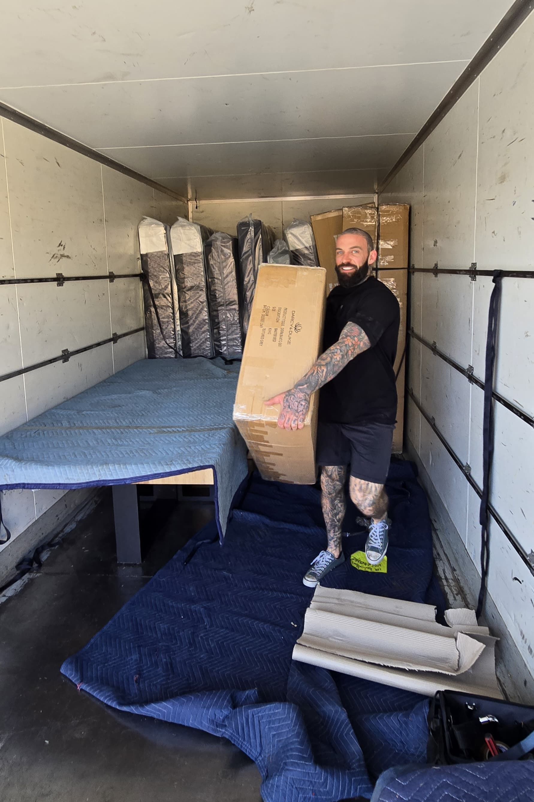 Man carrying a large cardboard box inside a truck, surrounded by moving blankets and furniture.