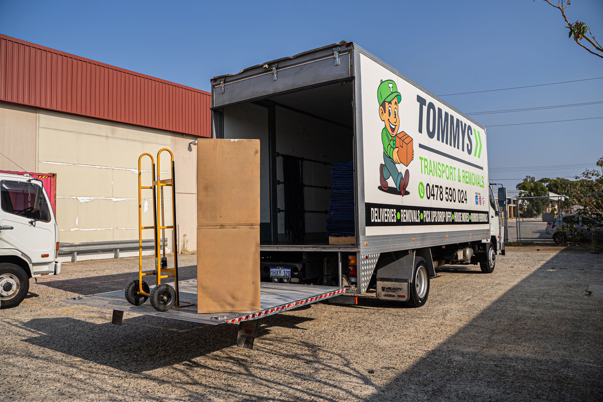 Box truck with open rear ramp parked beside a building, displaying a TOMMY'S - Transport and Removal