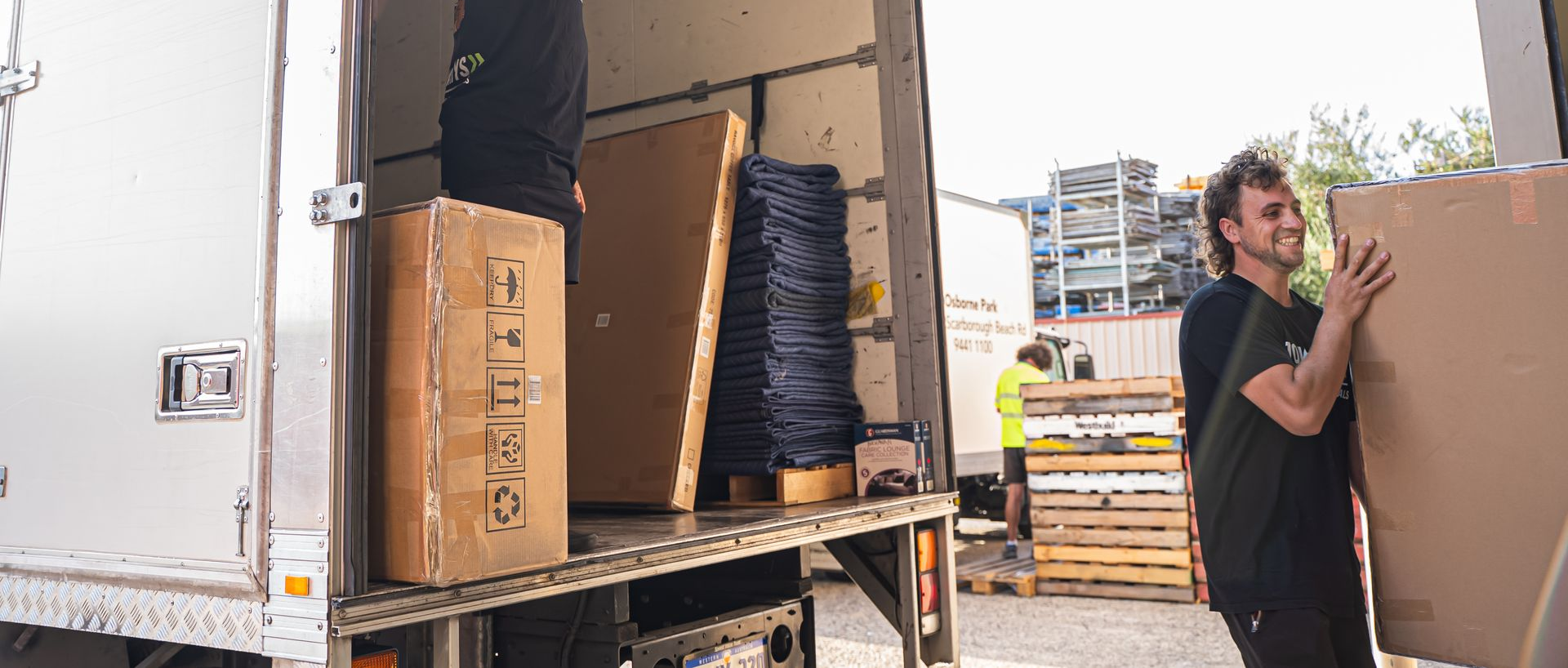 Removalist carrying a large cardboard box beside a moving truck and stacked pallets in a warehouse yard.