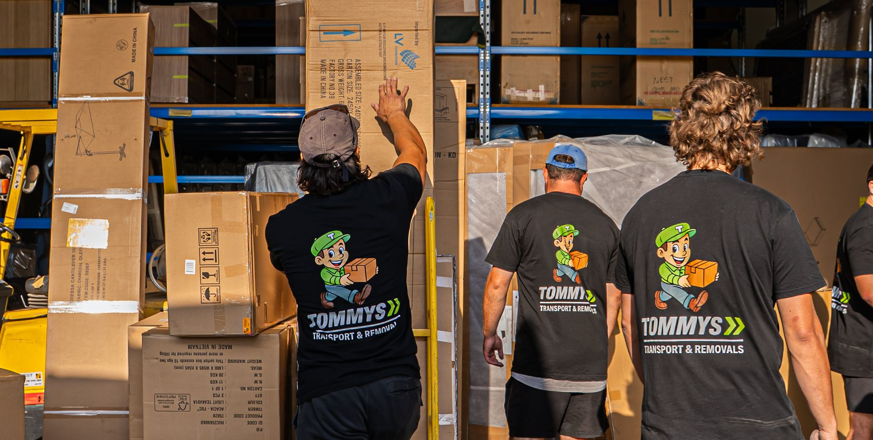 Removalist in black Tommy's removalist T-shirts stacking boxes in a warehouse.