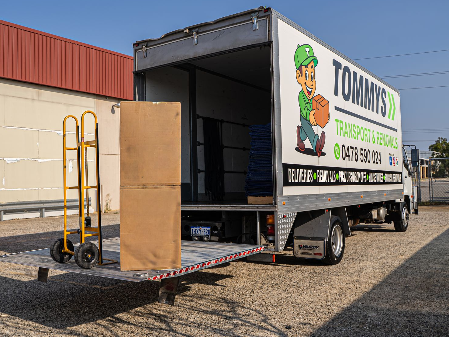 Moving truck with open back and TOMMY’S Removalist logo on the the side of the truck which is parked beside a building.