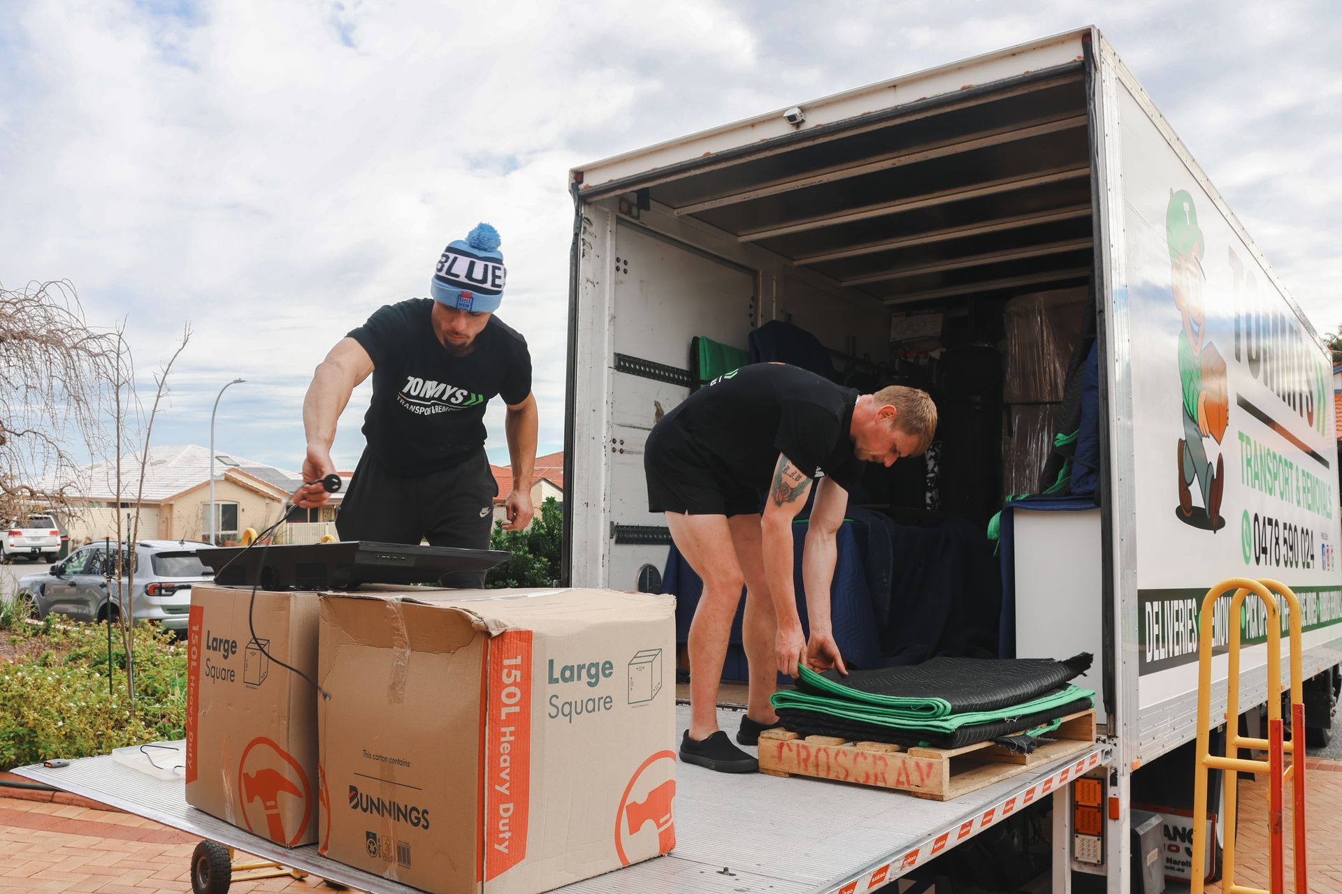 Two workers from Tommy's loading boxes and a pallet into a delivery truck outdoors.