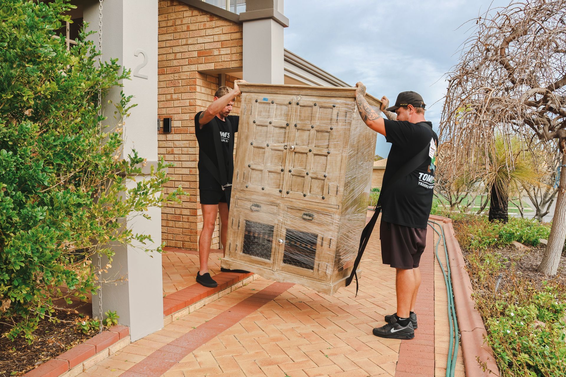 Two workers carrying a large appliance through a brick walkway outside a house