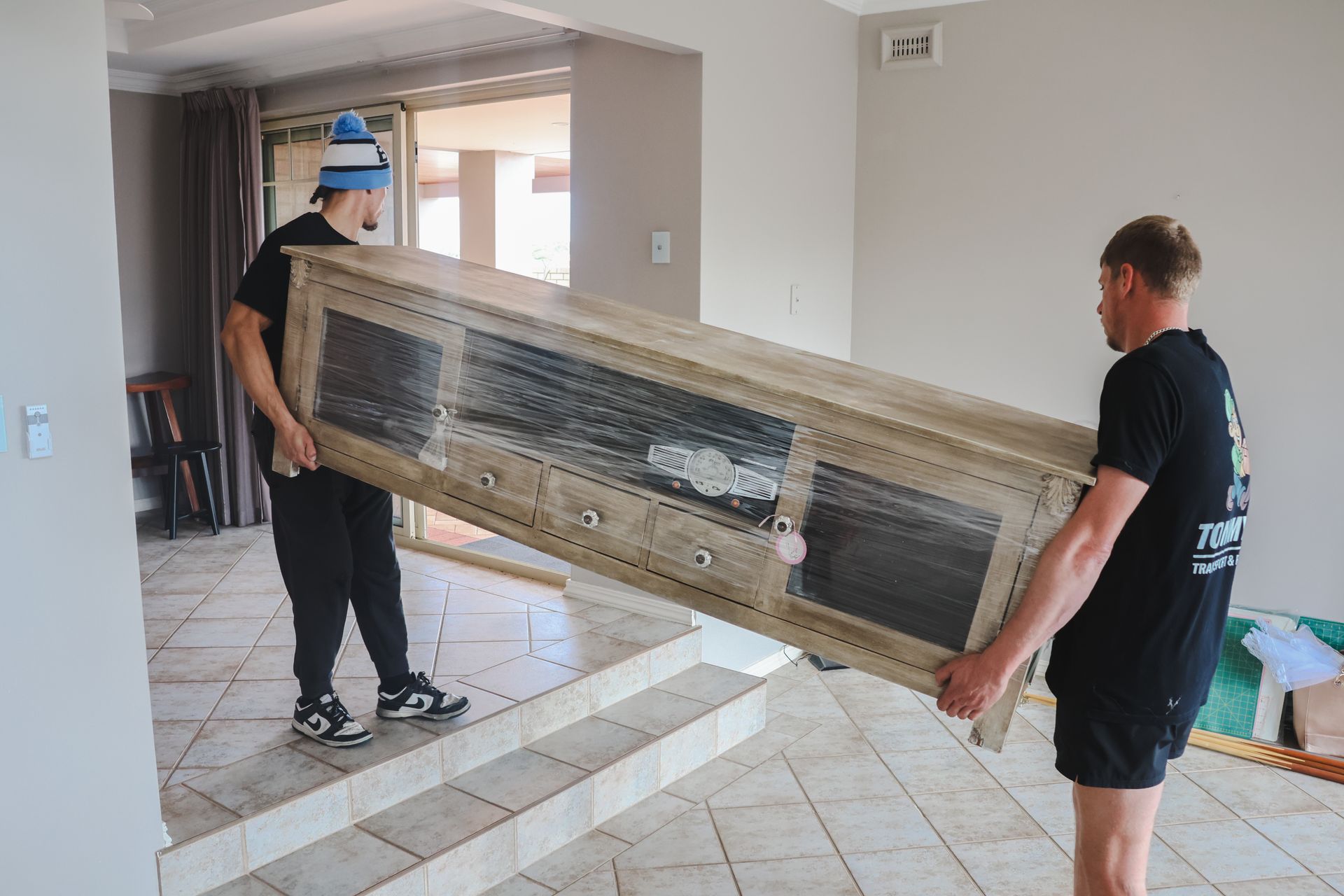Two of Tommy's workers carrying a large wrapped TV cabine through a doorway in a tiled home interior