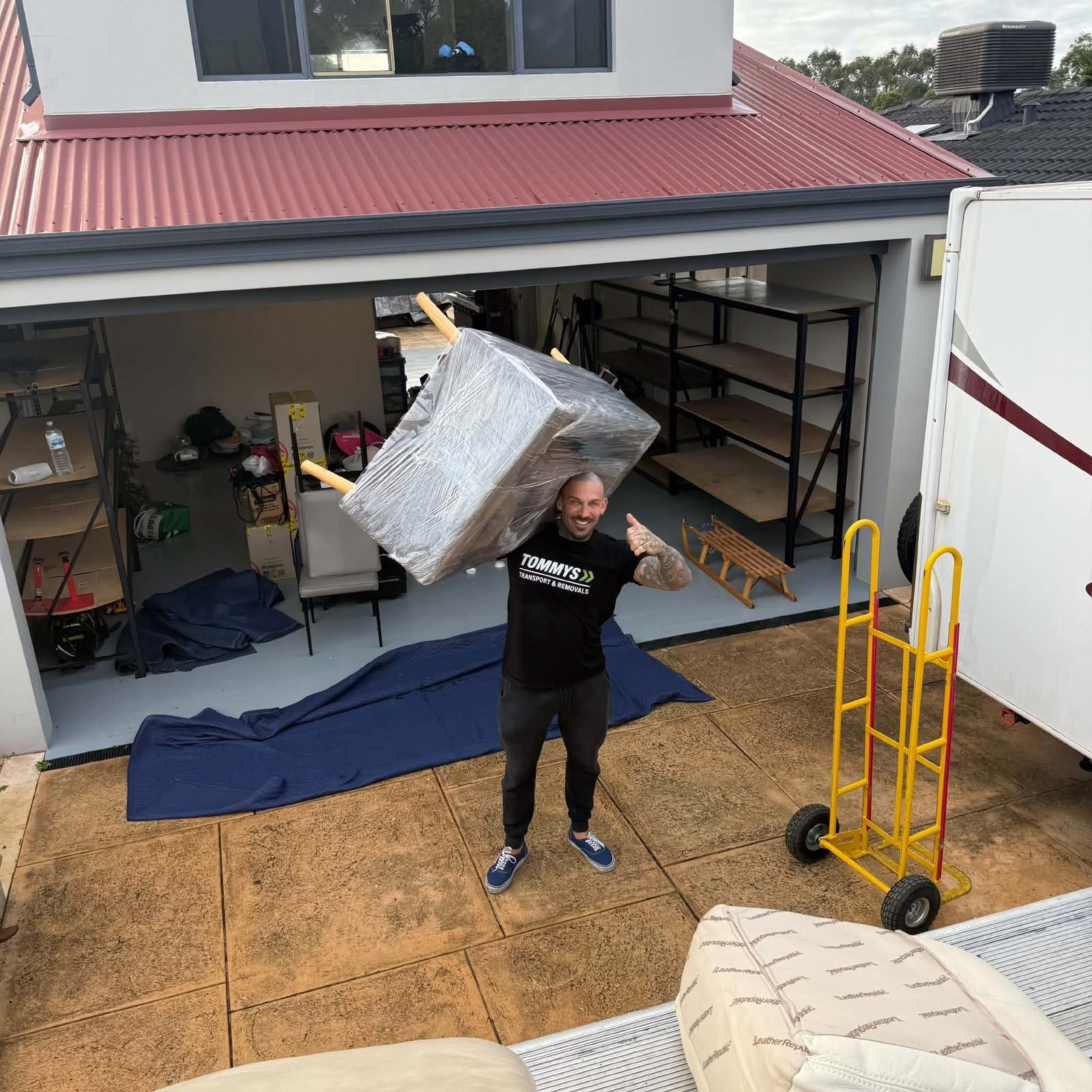 Person in a garage lifting a large wrapped item overhead, with moving blankets and hand truck nearby.