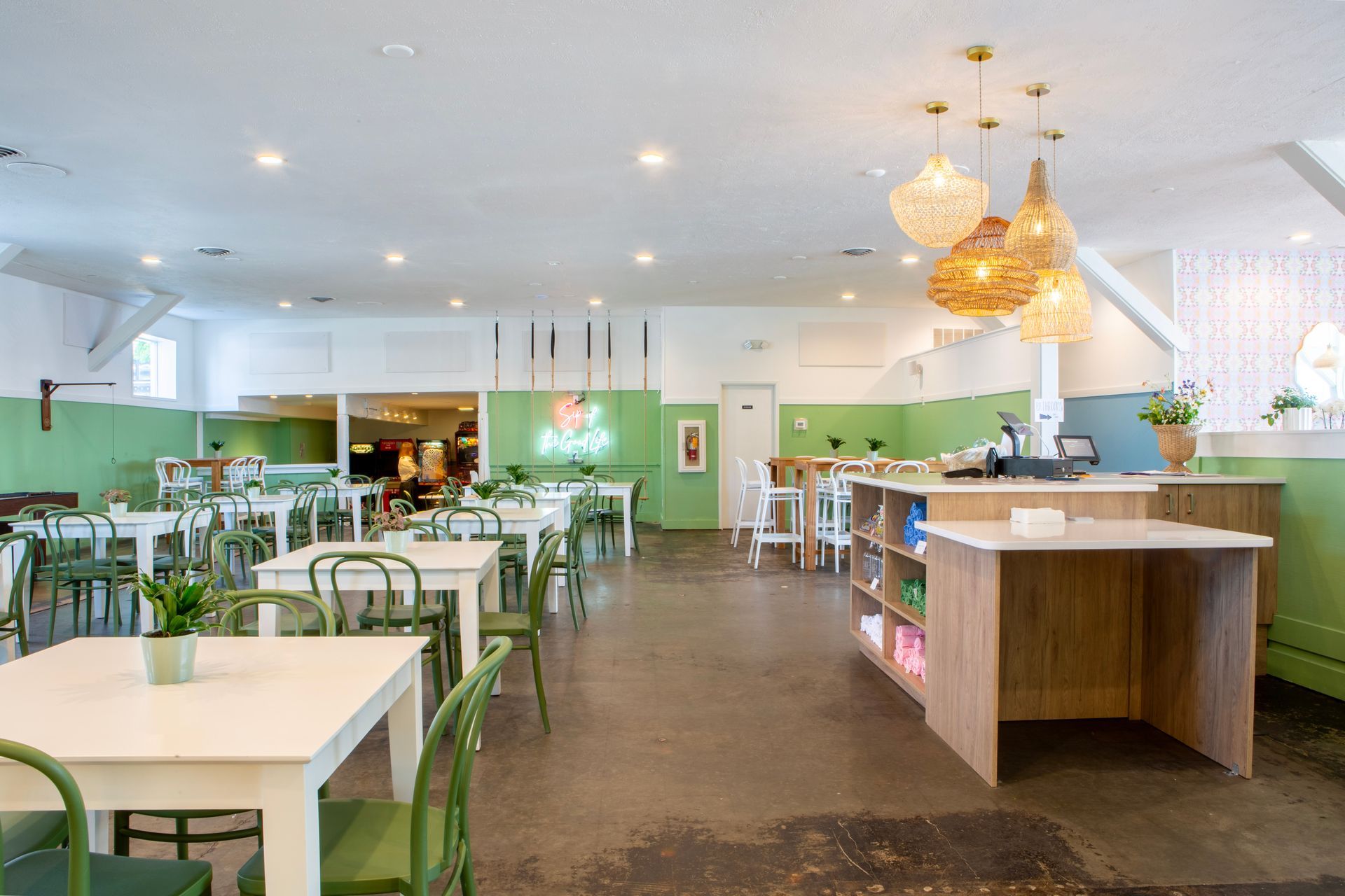 Bright, airy cafe interior with white tables, green chairs, and light green accents. Counter and hanging lamps visible.