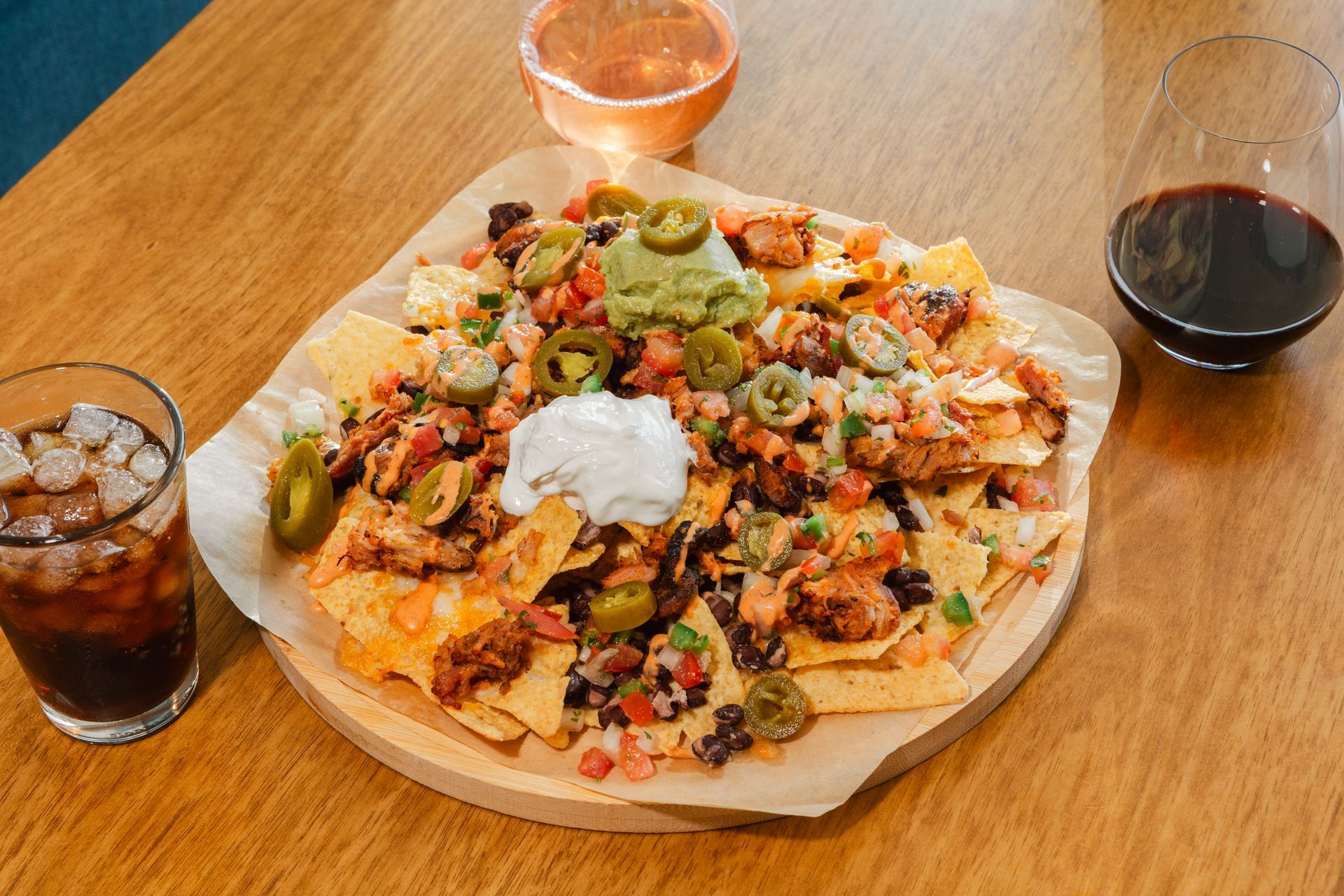 Nachos with toppings like guacamole, sour cream, and jalapeños on a wooden serving board, with drinks.