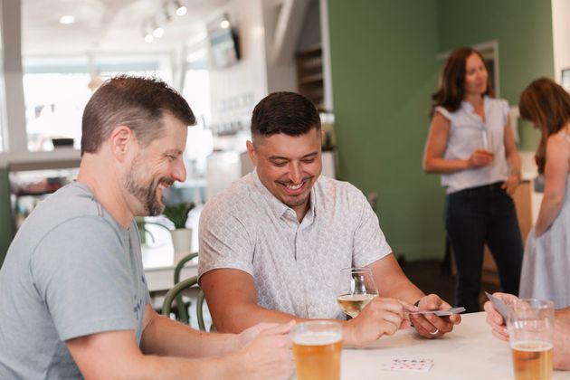 Two men playing cards and smiling at a table in a bar; two women stand nearby.