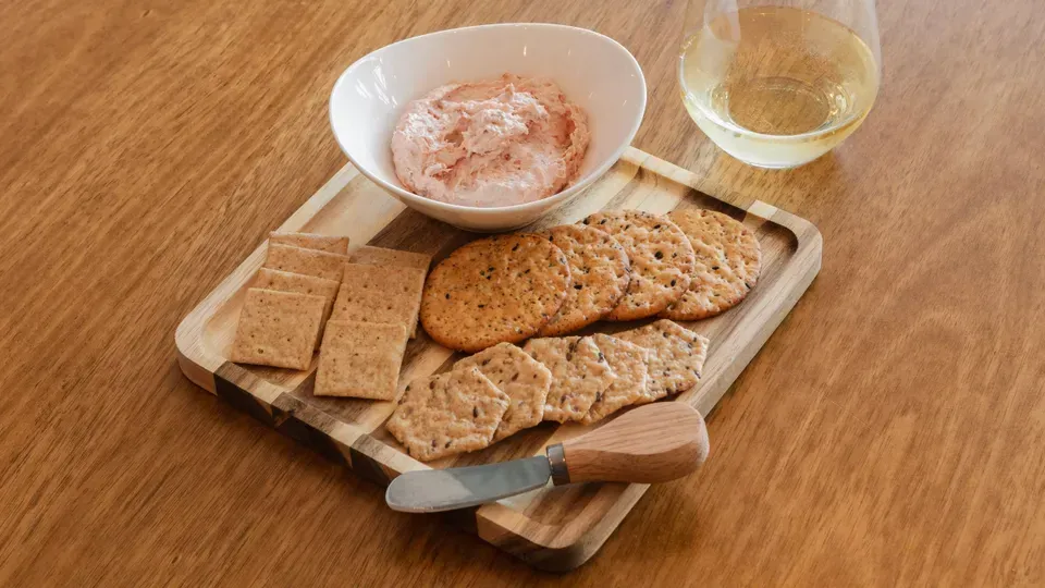 Wooden serving board with crackers, pink dip, knife, and a glass of white wine.