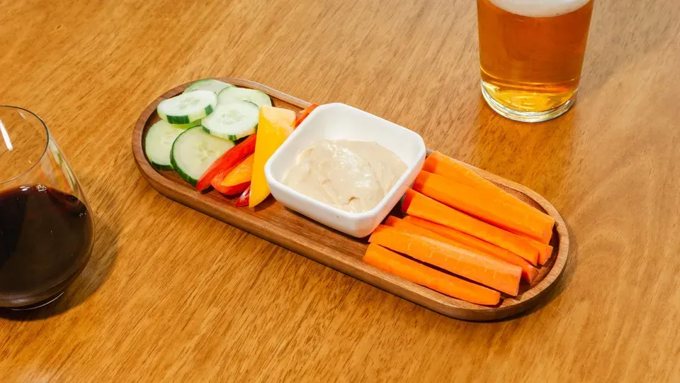 Wooden platter with vegetables and dip, accompanied by drinks on a table.