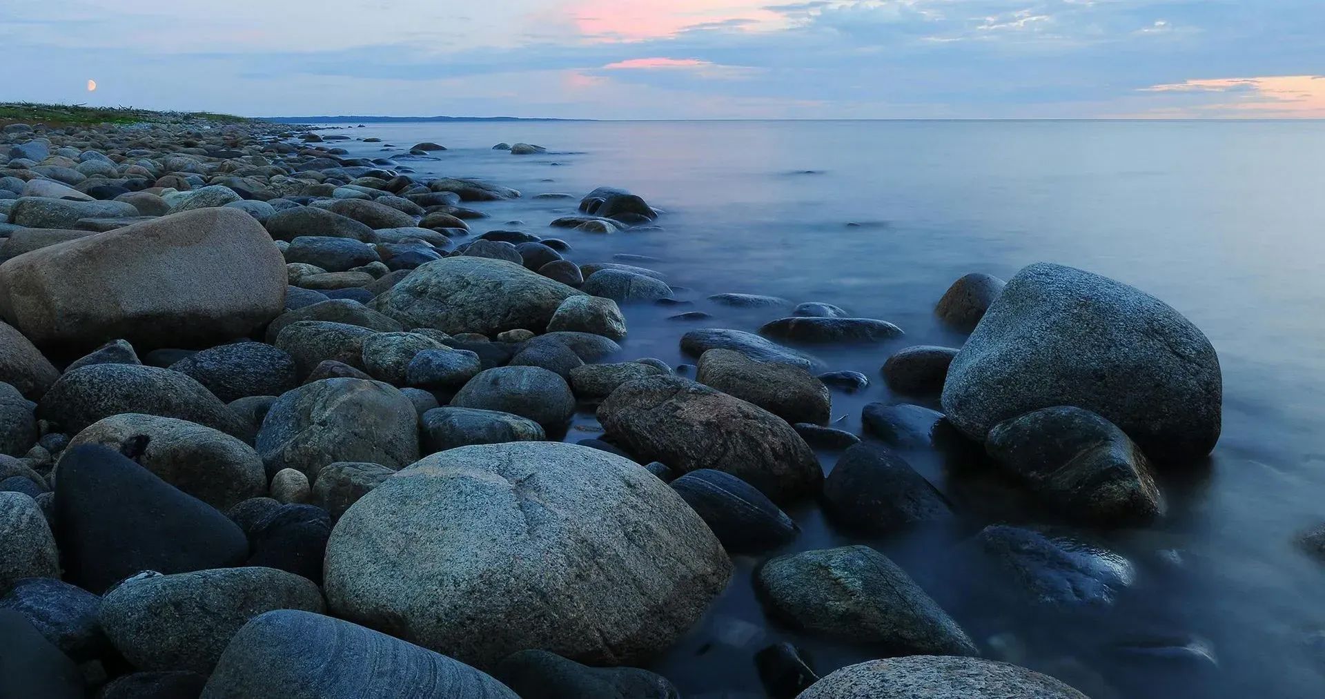 A rocky shoreline leads into a calm, dark sea under a soft, twilight sky with faint pink clouds.