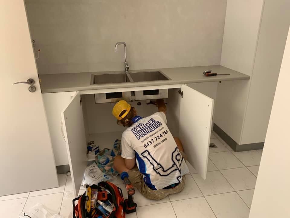 Plumber Installing Pipes Under a Kitchen Sink in a White Cabinet — Langley Plumbing in Mareeba, QLD
