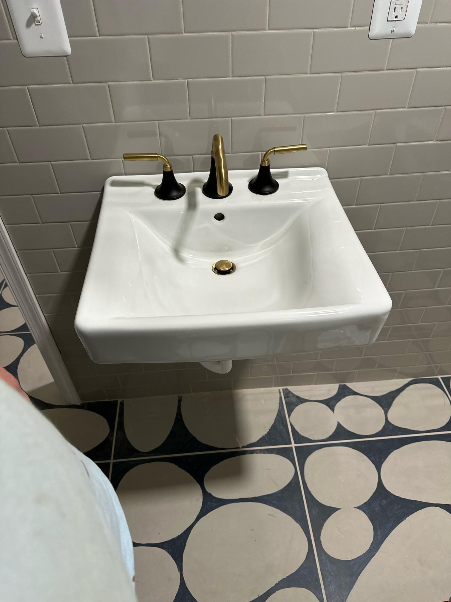 White wall-mounted sink with gold and black fixtures against gray tile and patterned floor.
