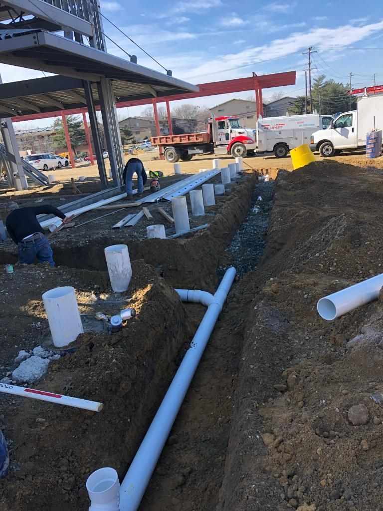 Construction site: workers installing pipes in a trench near a building, under a blue sky.