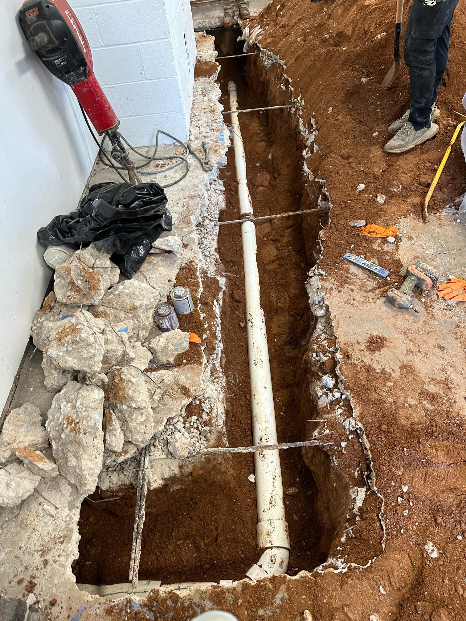 Trench dug along a wall, exposing white pipes; tools and dirt pile nearby, with a person's feet visible.